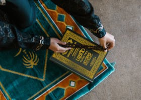 Top view of a person holding prayer beads and Quran on a colorful rug, symbolizing Islamic faith and worship.